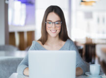 Portrait of cheerful Caucasian female student wearing eyeglasses using laptop in cafe for studies or networking and smiling